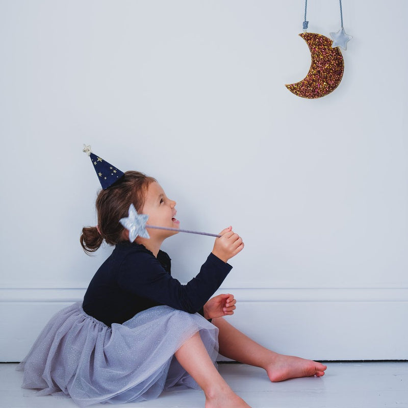 girl sits on the floor with a want and dress up hat looking at the moon in the grey sparkle gold glitter dot and gold stars tutu by Mimi and Lula and Crane and Kind 